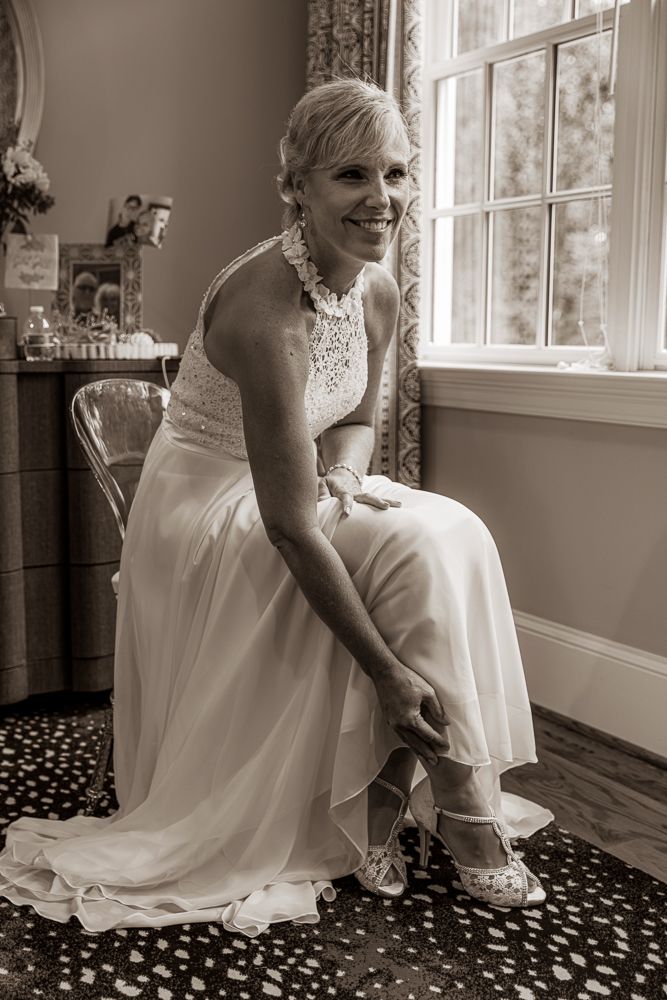 Bride in white halter gown adjusting her heels while seated by a window in sepia-toned wedding photo.