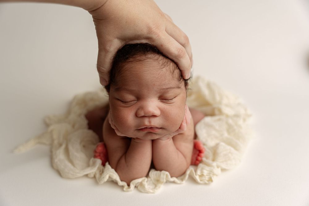 Sleeping newborn baby posed with hands under chin on white blanket, with parent's hand gently cradling head.