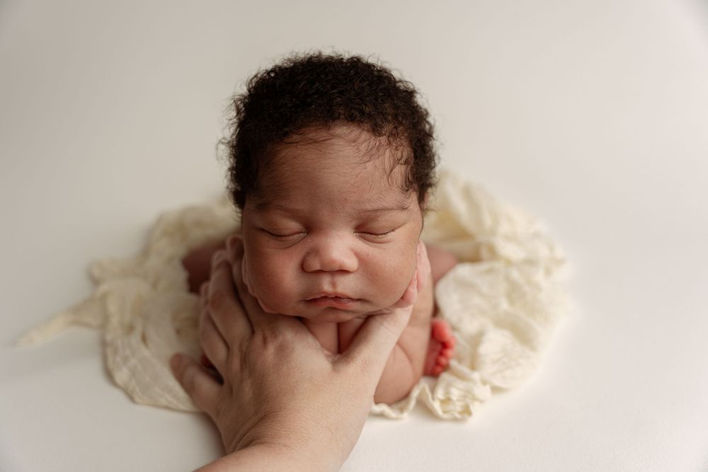 Peaceful newborn baby sleeping with chin resting on hands, nestled in cream fabric during professional newborn photography session.