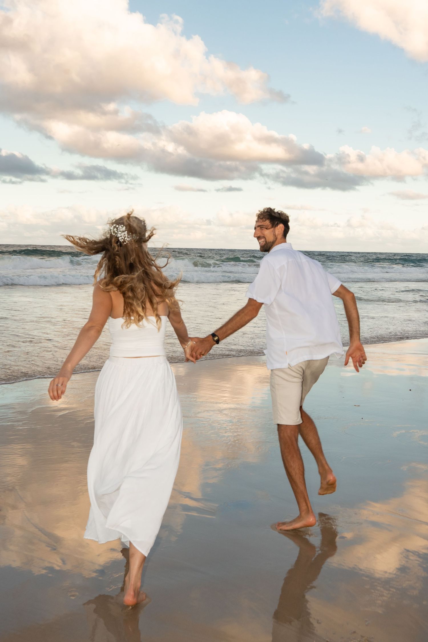 Couple holding hands and running on beach at sunset in white summer outfits with waves and clouds in background.