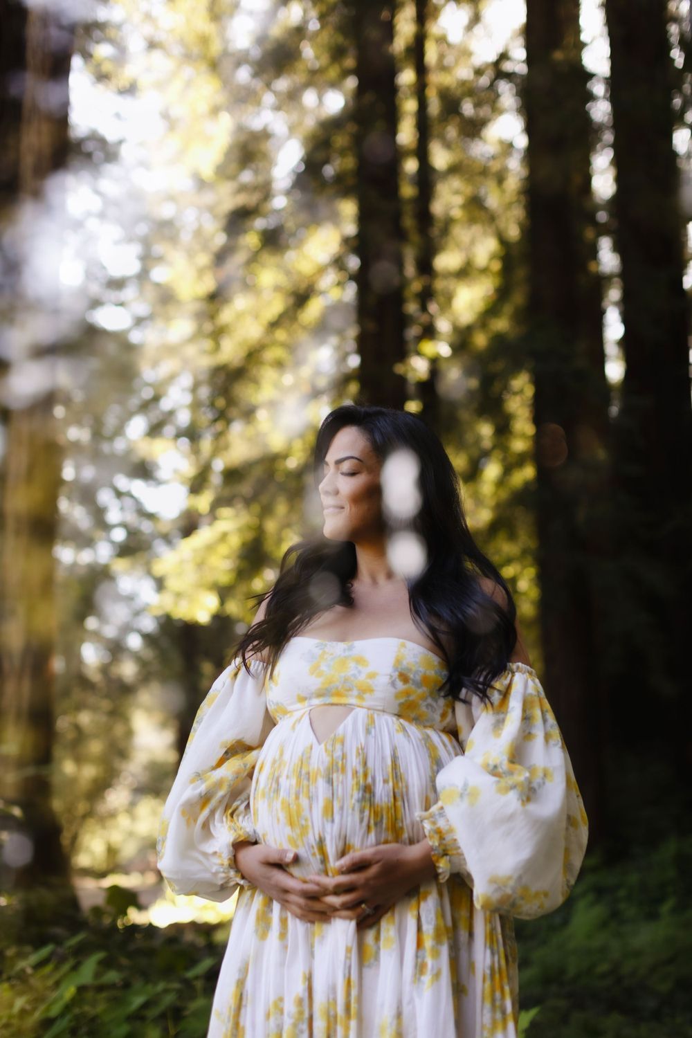 Double exposure of expectant mother in white and yellow floral gown poses with hand on baby bump by pine trees with sunlit bokeh.