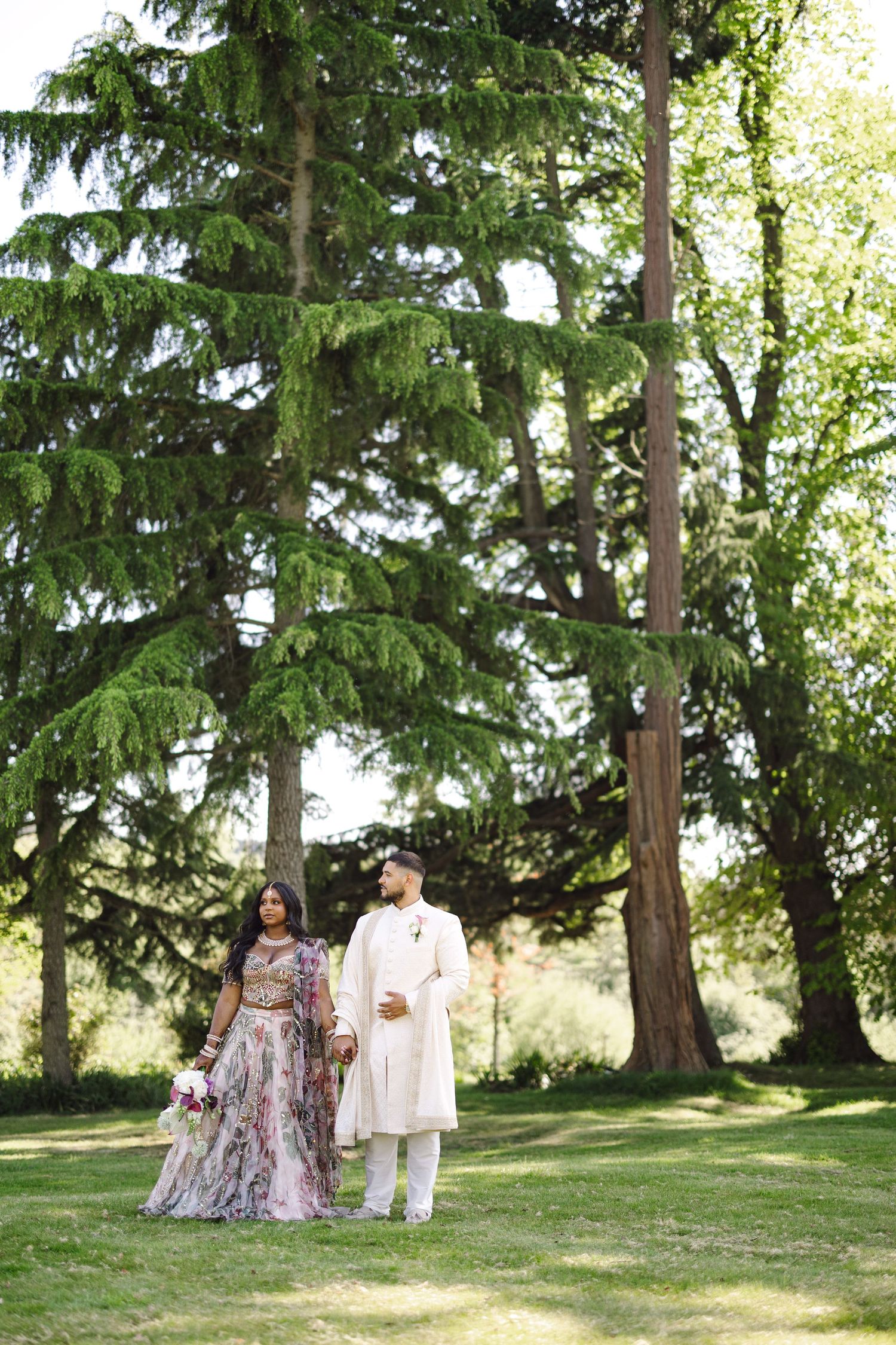 A couple walks together on a grassy lawn surrounded by tall evergreen trees on a sunny summer day.
