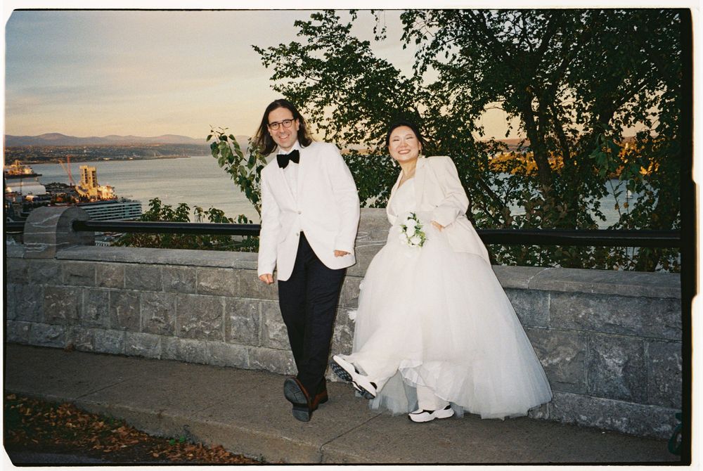 Bride and groom posing outdoors near a stone wall with a scenic river and city view at dusk.