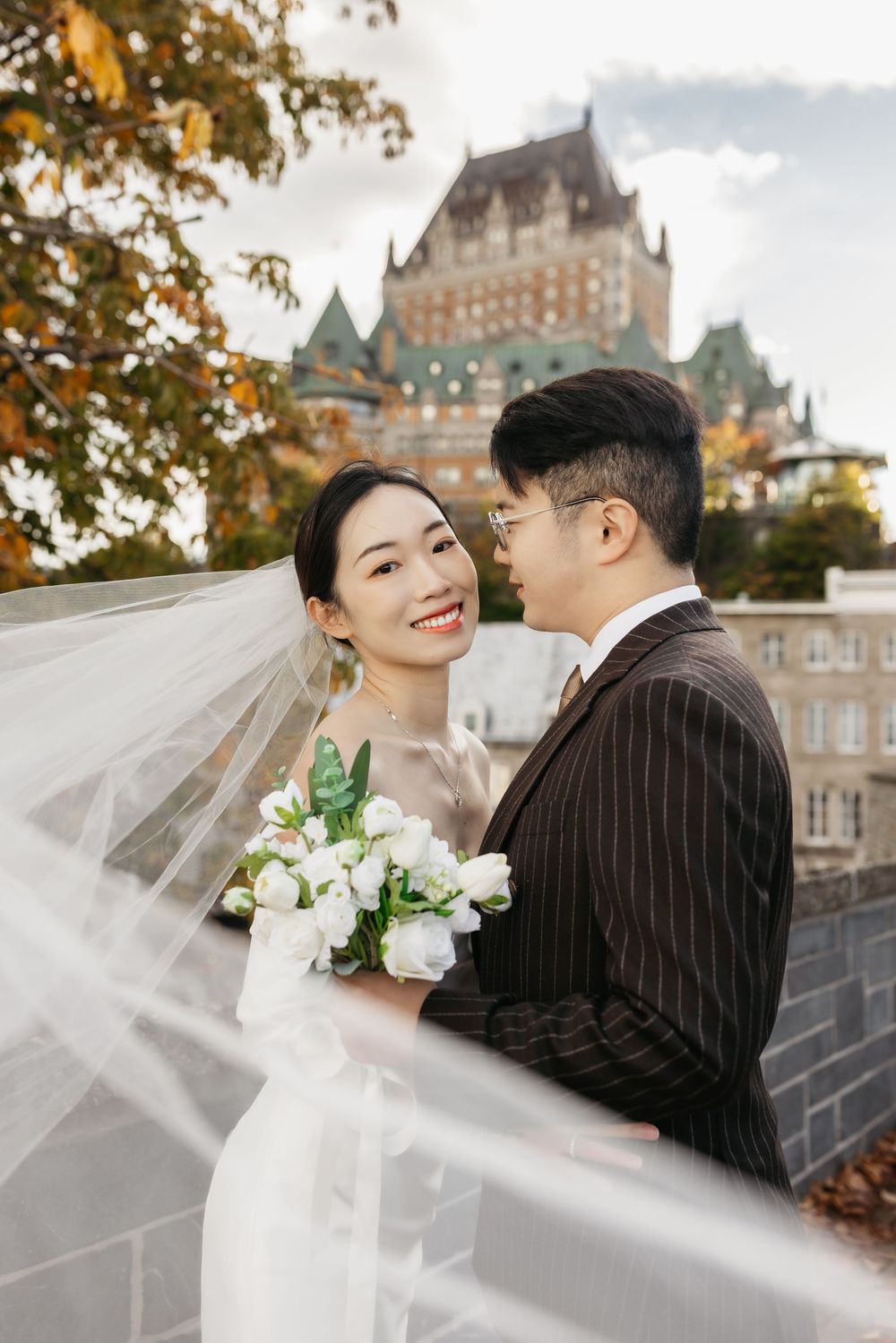 Asian couple in wedding attire poses before Château Frontenac in Quebec City, bride holding white roses bouquet.