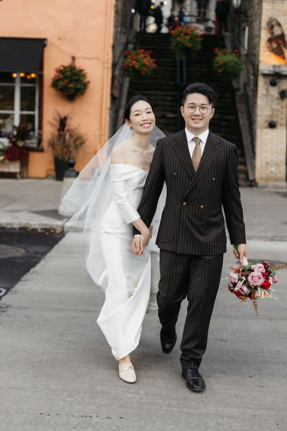 A smiling Asian bride in a white off-shoulder gown and veil walks with a groom in a pinstripe suit holding a bouquet.
