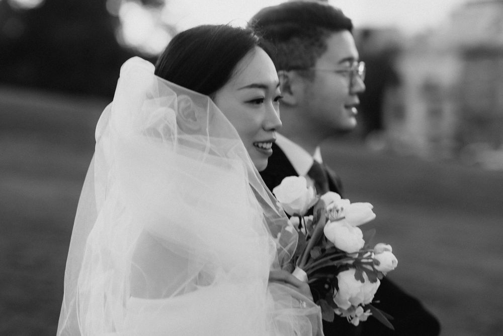 Black and white wedding photo of a smiling Asian bride holding white flowers beside her groom outdoors.
