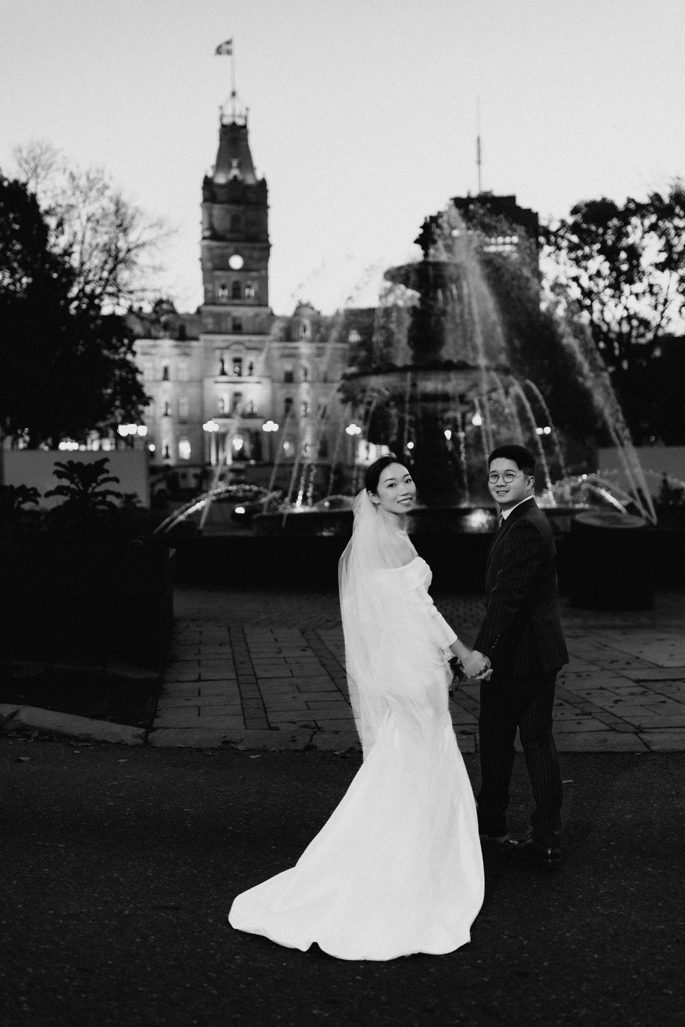 Black and white wedding photo of bride and groom holding hands before a fountain and historic clock tower at dusk.