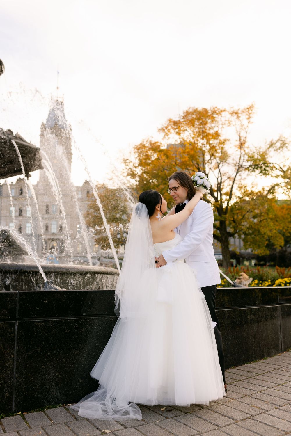 Bride and groom embracing near a fountain with autumn trees in the background on their wedding day.