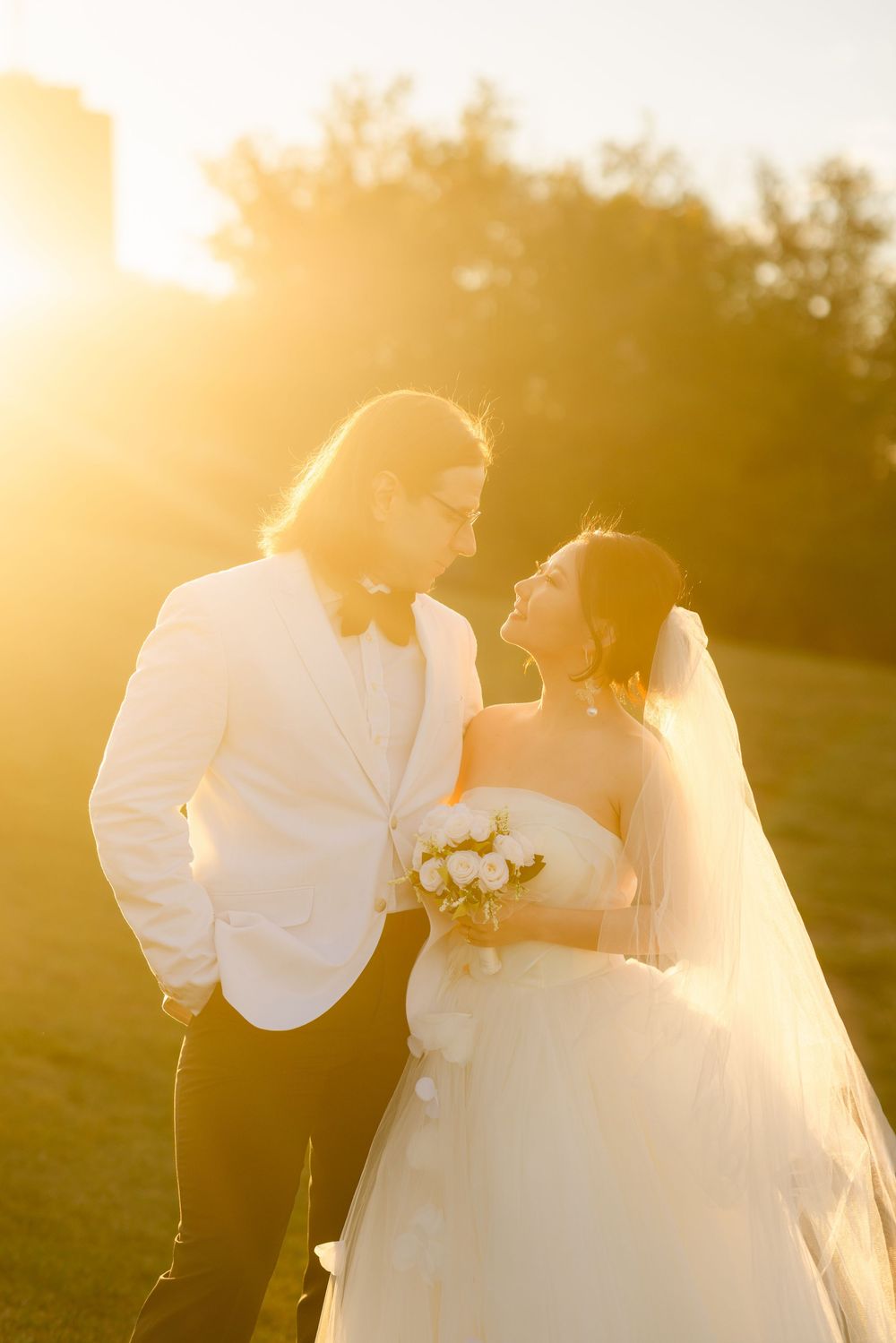 Bride and groom share a romantic moment outdoors, bathed in golden sunset light, as she holds a white bouquet.