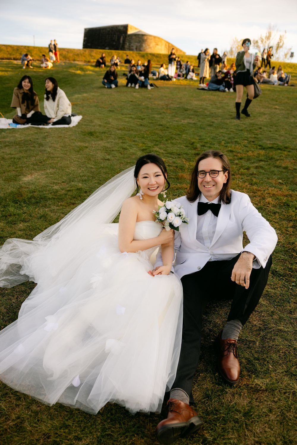 Bride and groom sitting on grass in wedding attire, white gown and tuxedo, holding bouquet outdoors.