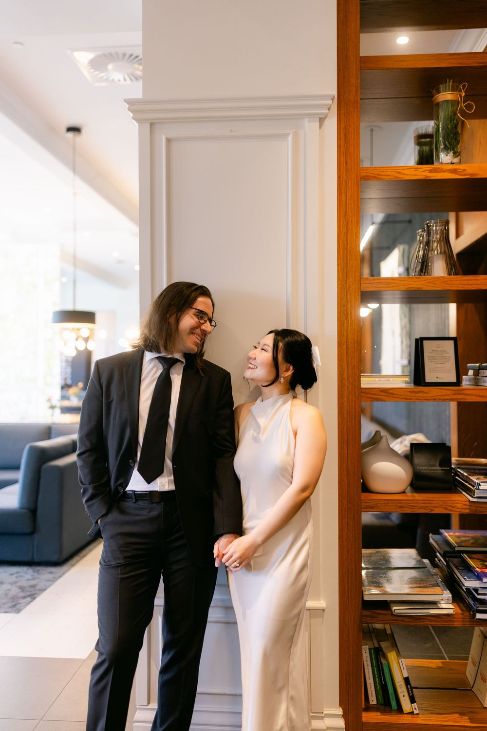 Elegant couple in formal attire sharing a romantic moment beside a wooden bookshelf in a stylish interior.