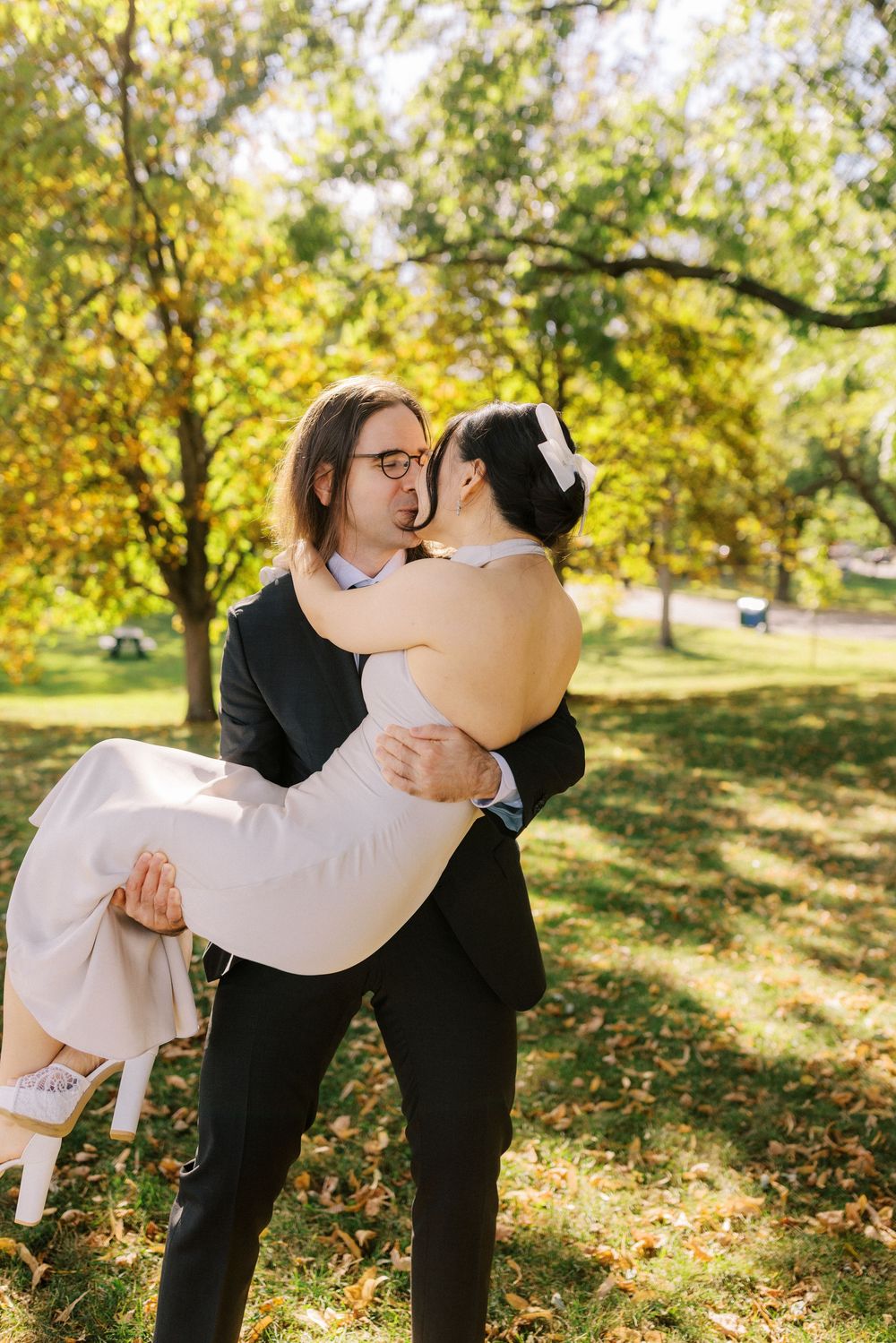 Groom in black suit carries bride in white dress while they kiss in a sunlit autumn park.