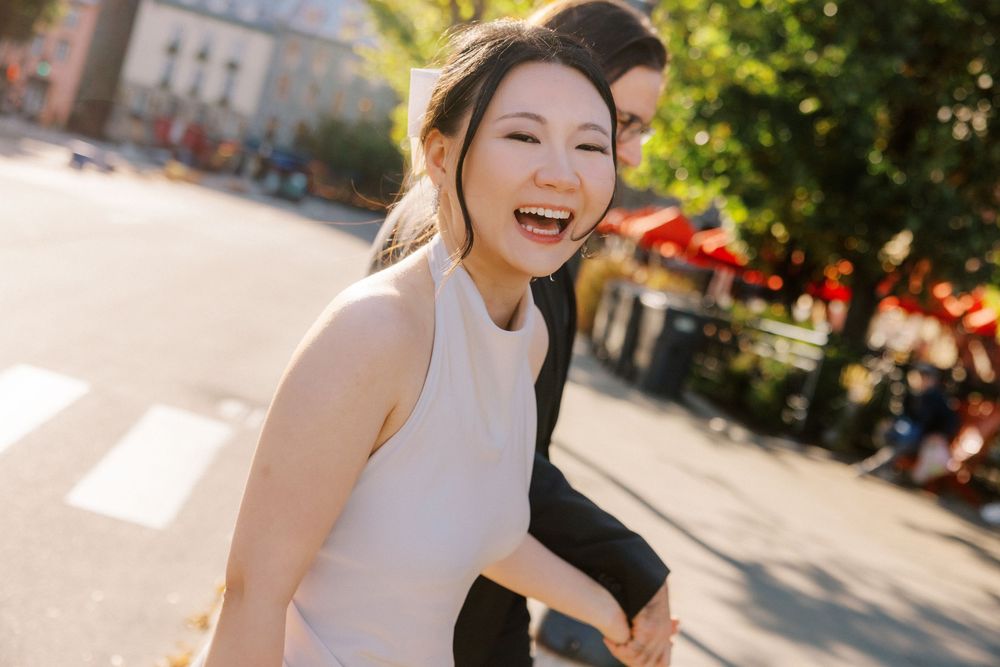 A joyful Asian woman laughs while holding hands with her partner on a sunny urban street.