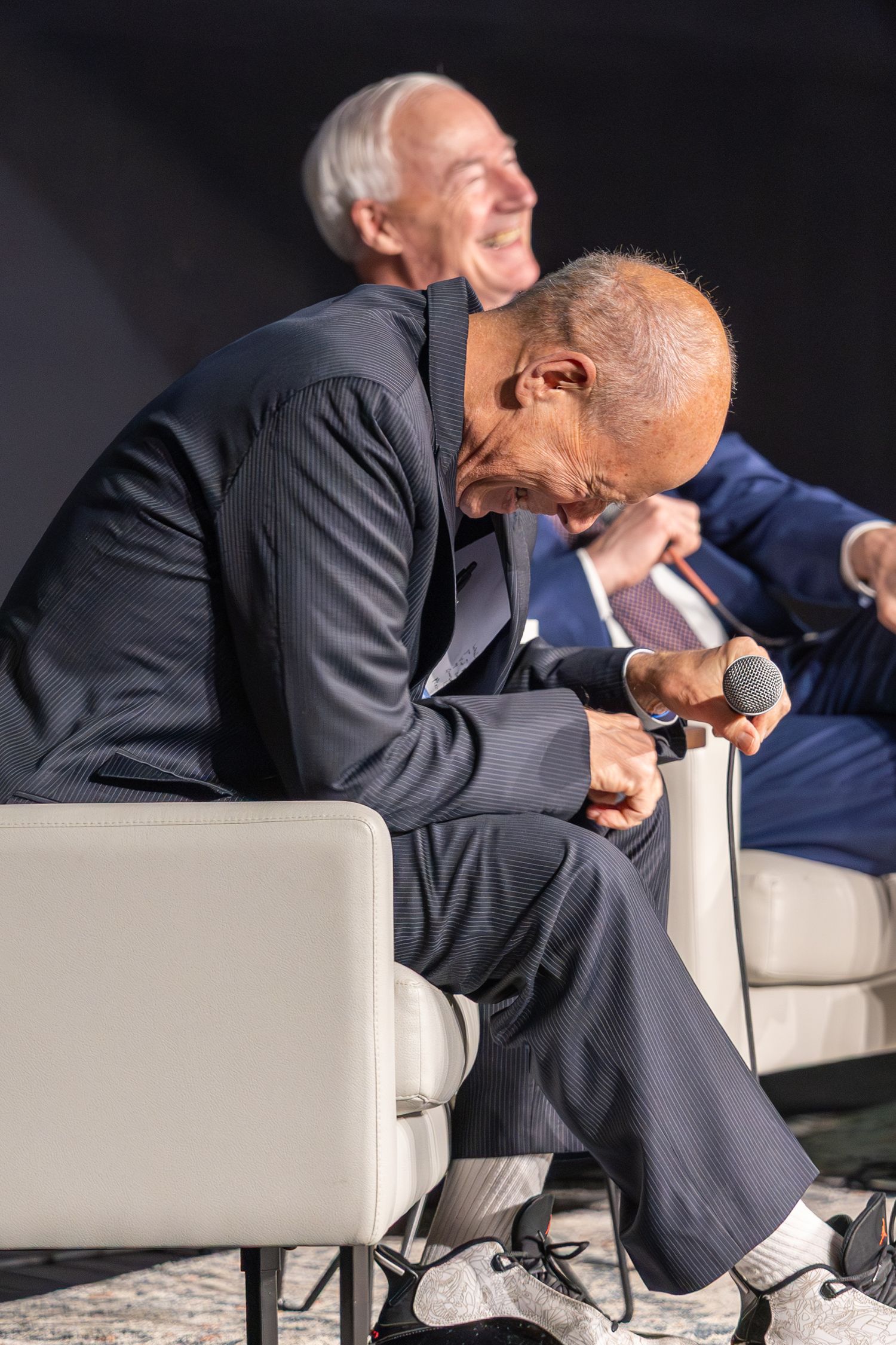 Two men in navy suits sit on white chairs at a panel event, one leaning forward laughing while holding a microphone.