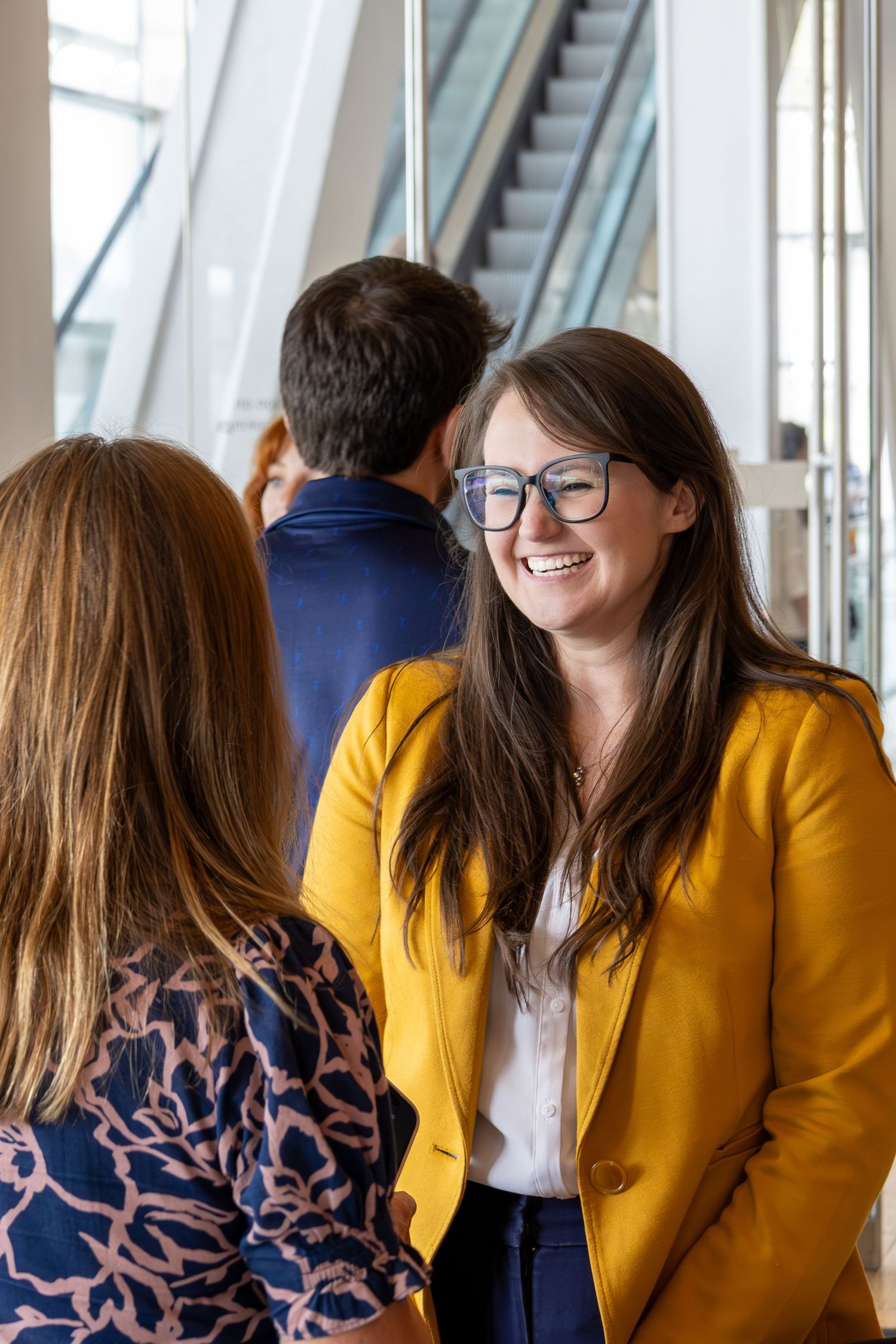 A smiling woman in a yellow blazer and glasses converses with colleagues in a modern building atrium.