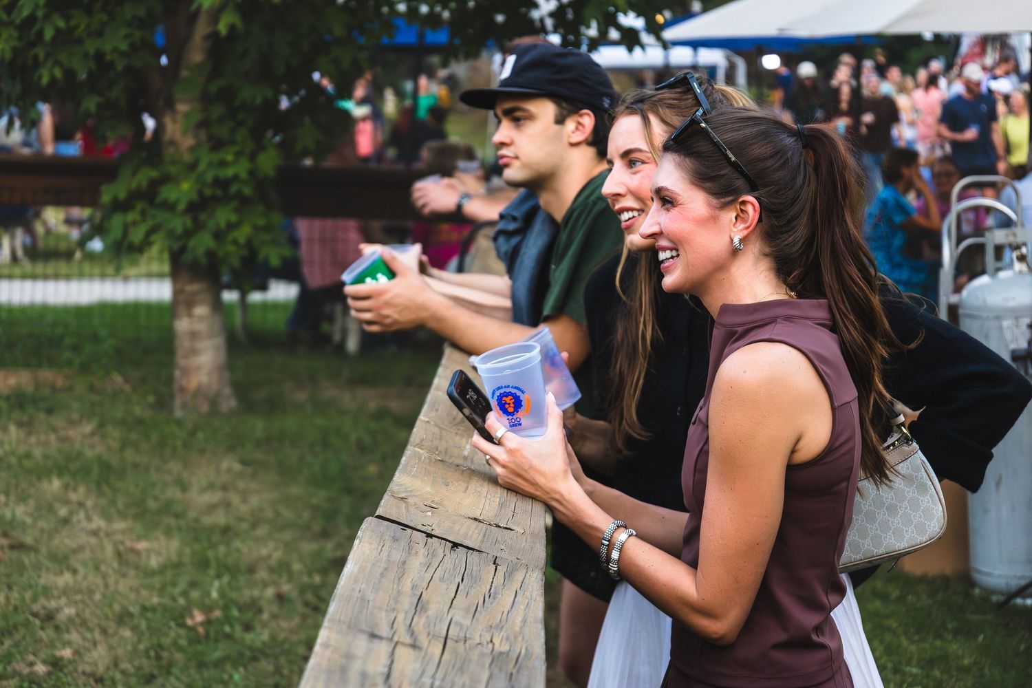 Two smiling friends enjoy drinks at an outdoor festival, standing near a wooden fence on a sunny day.