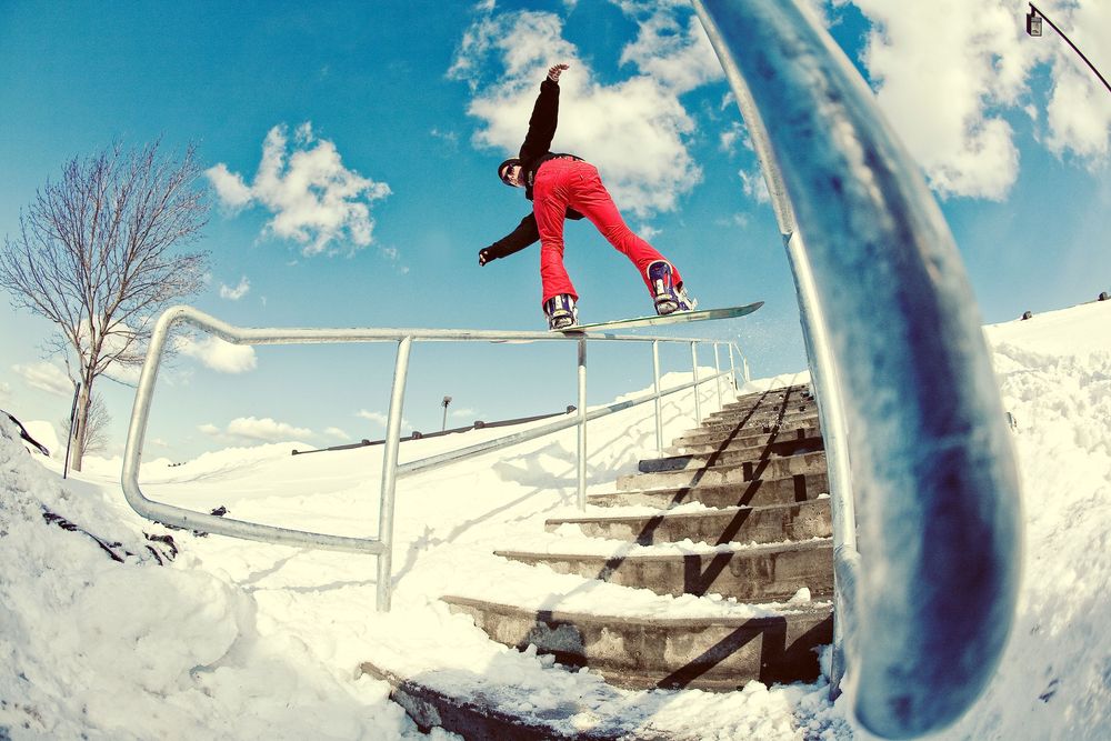 Snowboarder grinding a rail on snowy stairs under blue sky in red pants and black jacket.