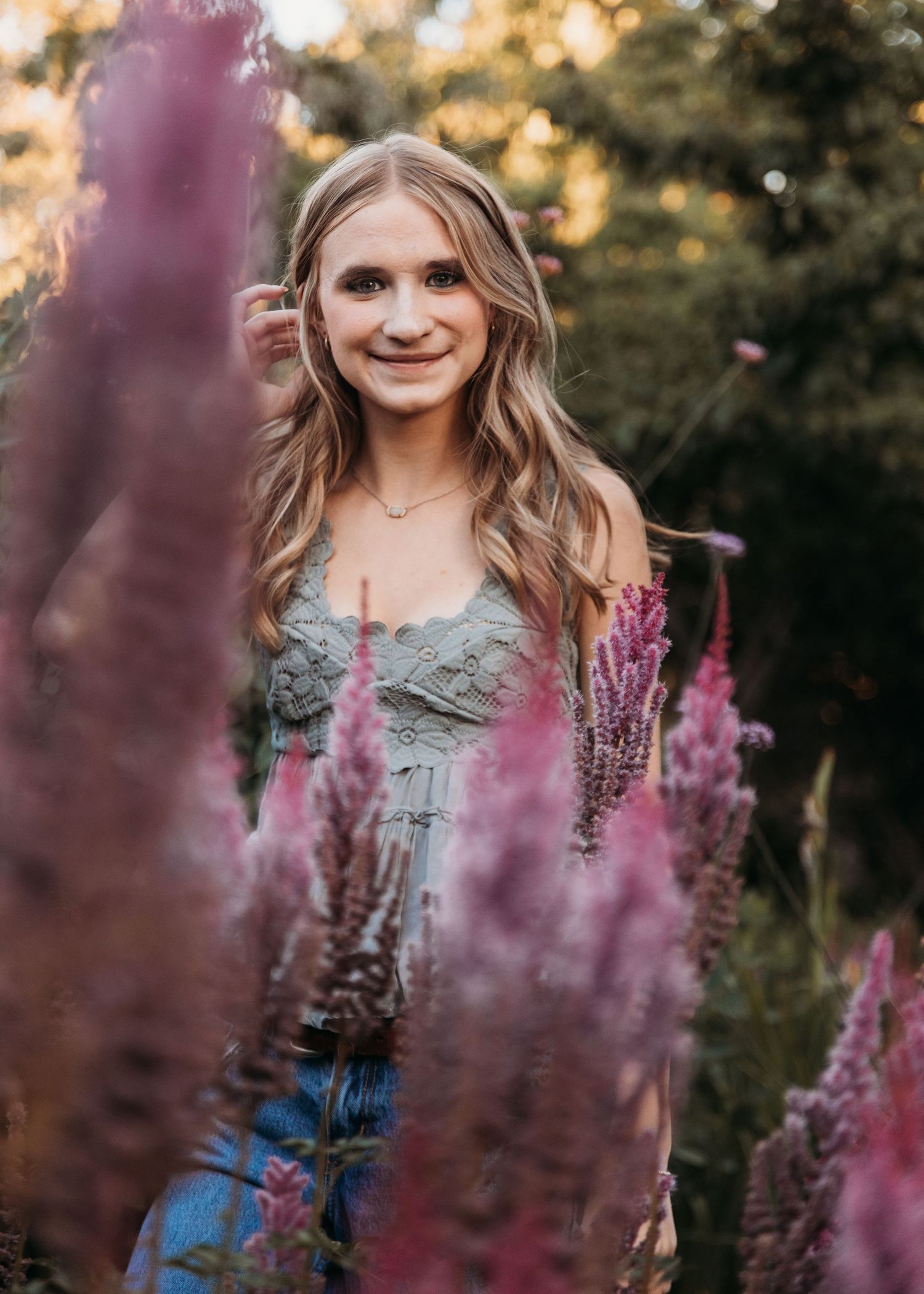 Young blonde woman smiling among pink wildflowers in a garden at golden hour, wearing a sage green lace dress.