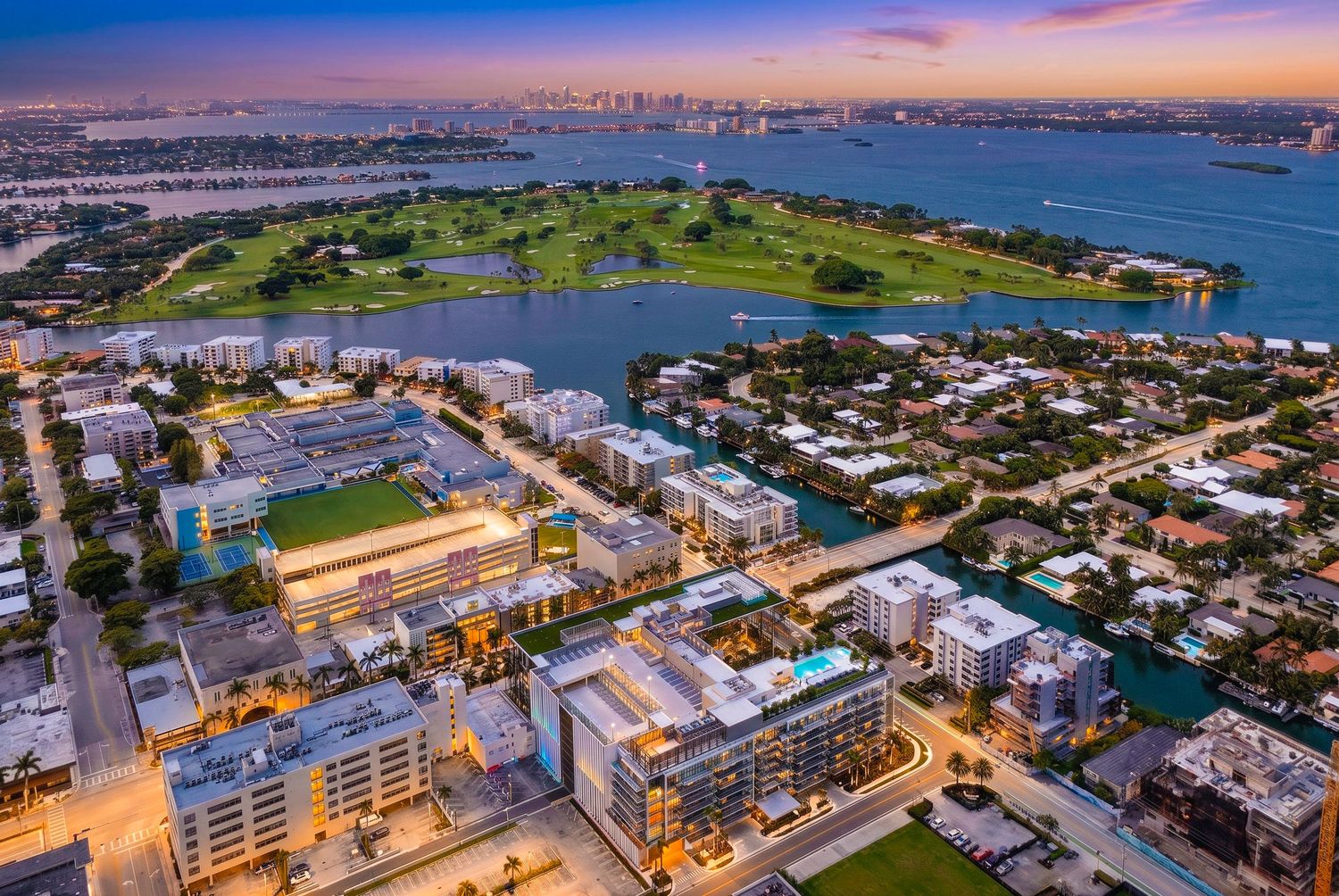 Aerial sunset view of Miami Beach waterfront buildings, lush green park, and city skyline reflecting on calm bay waters.