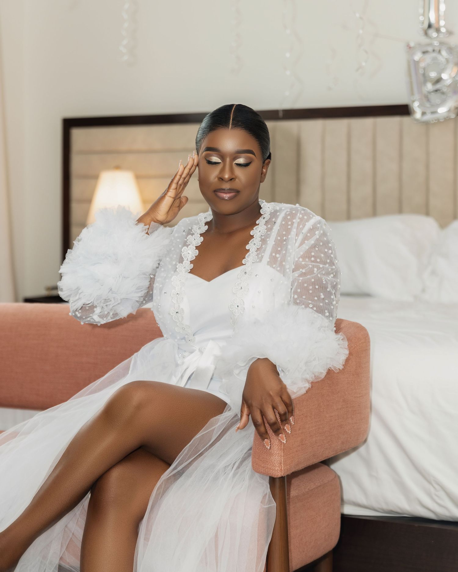 Bride in white ruffled tulle robe sits elegantly on pink chair in bridal suite.