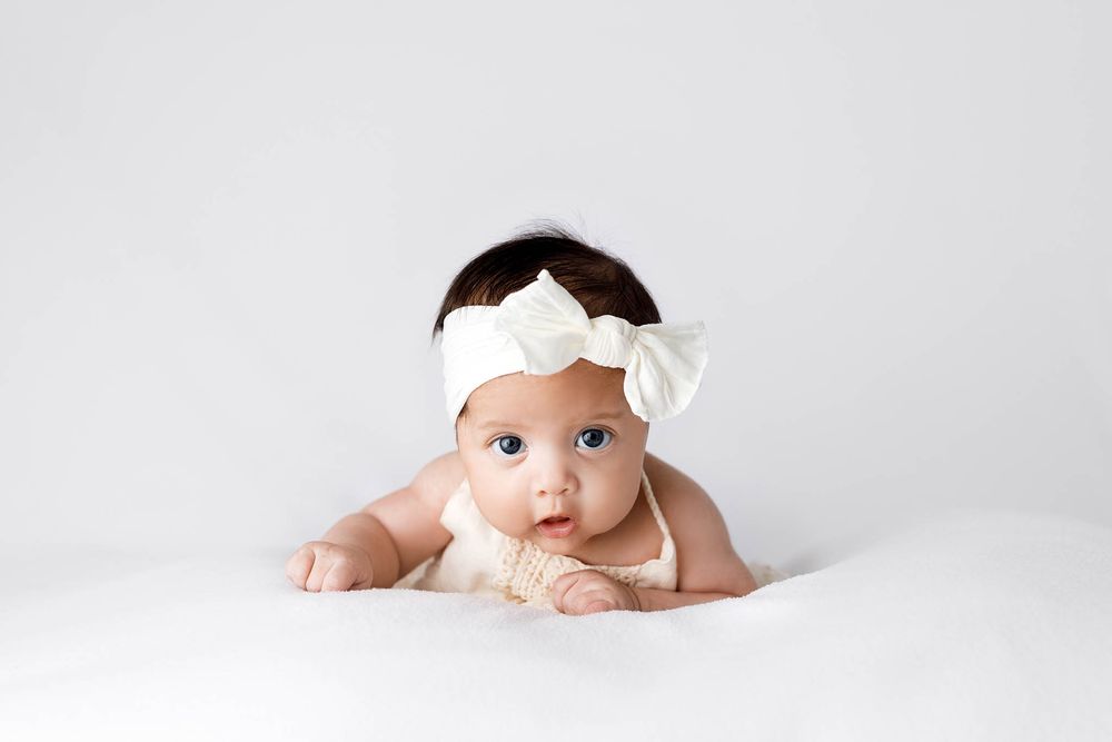 Adorable baby girl in white bow headband doing tummy time on white background.