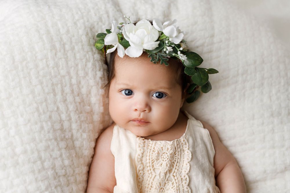Adorable baby girl wearing a white floral crown lying on a soft white blanket in a cream lace outfit.