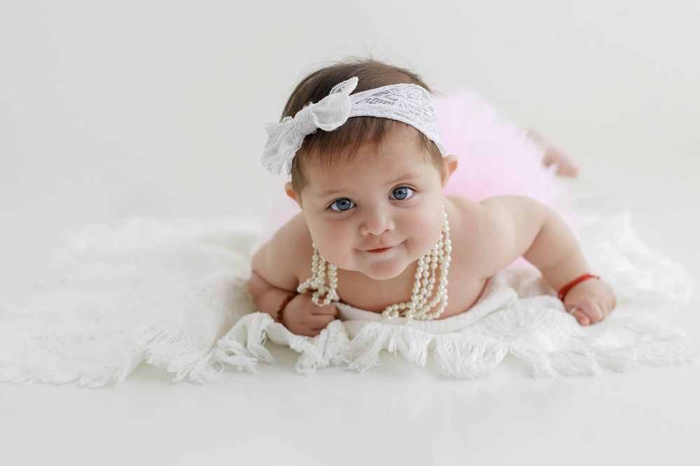 Baby girl in pink tutu and pearl necklace with lace headband poses on white blanket in studio photo.