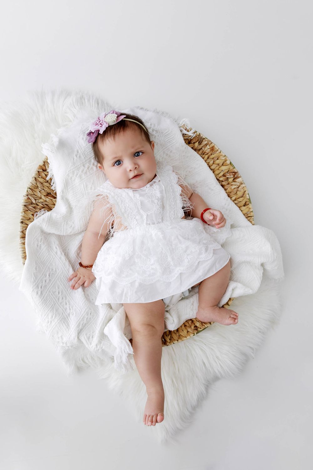 Baby girl in white lace dress and floral headband lying in wicker basket with fluffy white blanket.