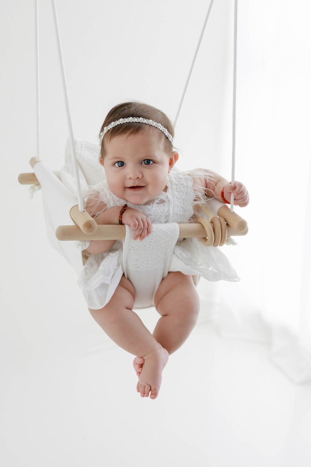 Adorable baby girl in white lace dress and headband sitting in a wooden swing against white background.