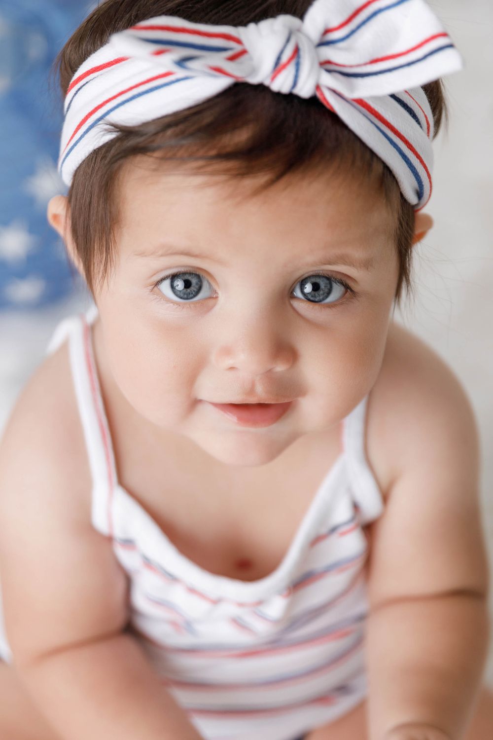 Cute baby girl with blue eyes wearing a striped headband bow and matching romper looking at camera.