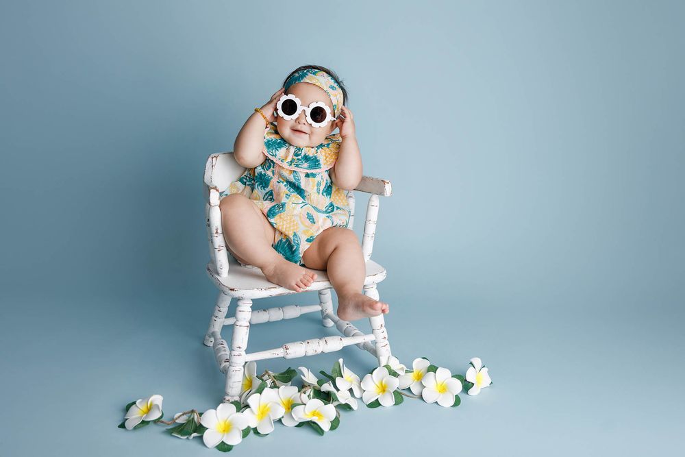 Stylish baby girl wearing sunglasses and floral romper sitting in a white wooden chair with flowers.