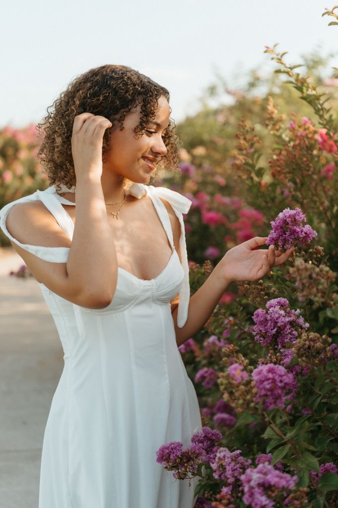 Smiling woman in white dress touching purple flowers in a blooming garden at sunset.