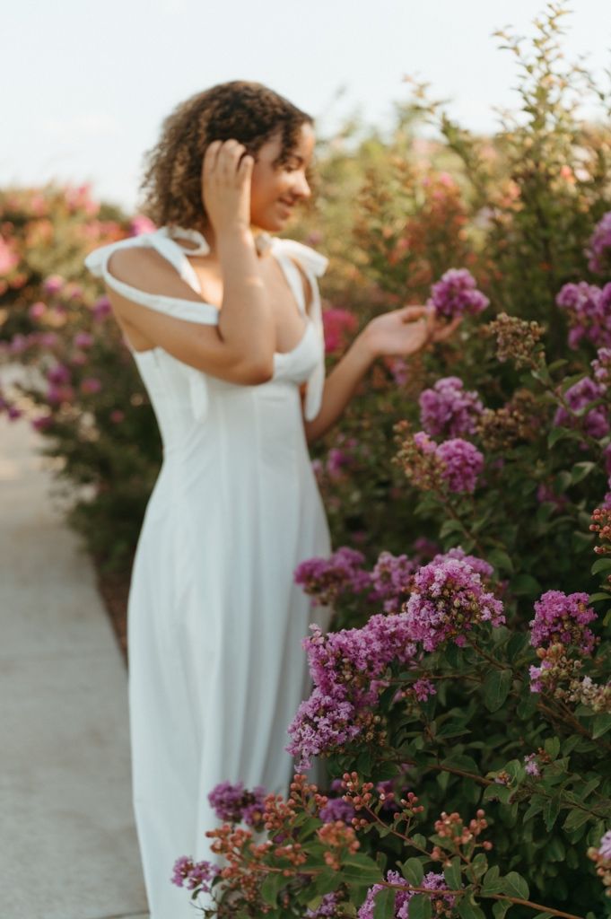 Woman in white dress smiling among purple flowering bushes in a garden at golden hour.