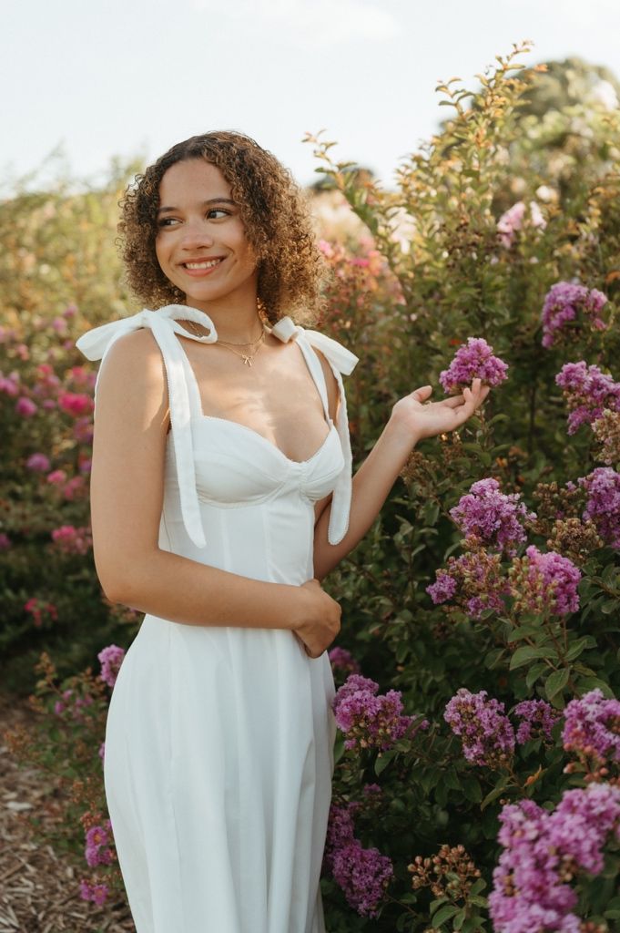Young woman in white sundress with bow straps smiling among pink flowering bushes in a garden.