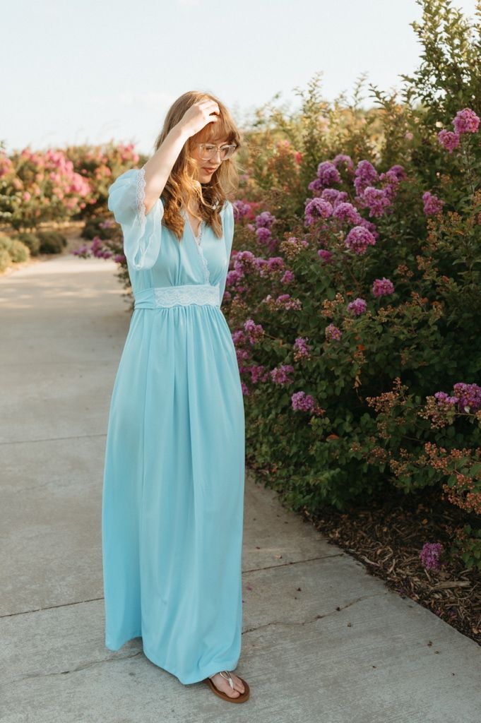 Young woman in light blue maxi dress poses on garden path with pink flowering bushes.