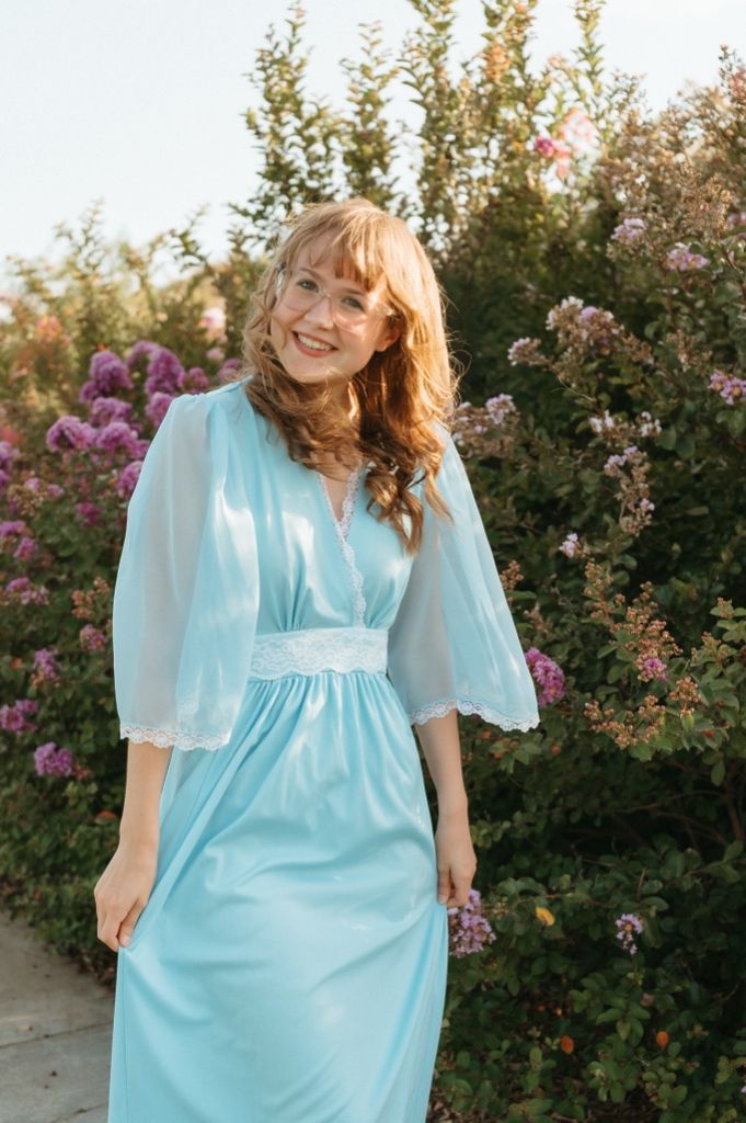 Woman in light blue maxi dress with lace waist and sheer sleeves smiling outdoors among blooming flowers.