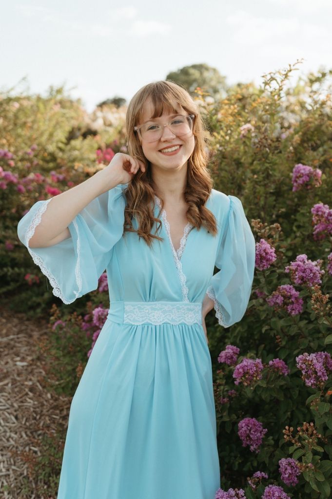 Smiling young woman in turquoise lace-trimmed dress stands among pink blooming shrubs.