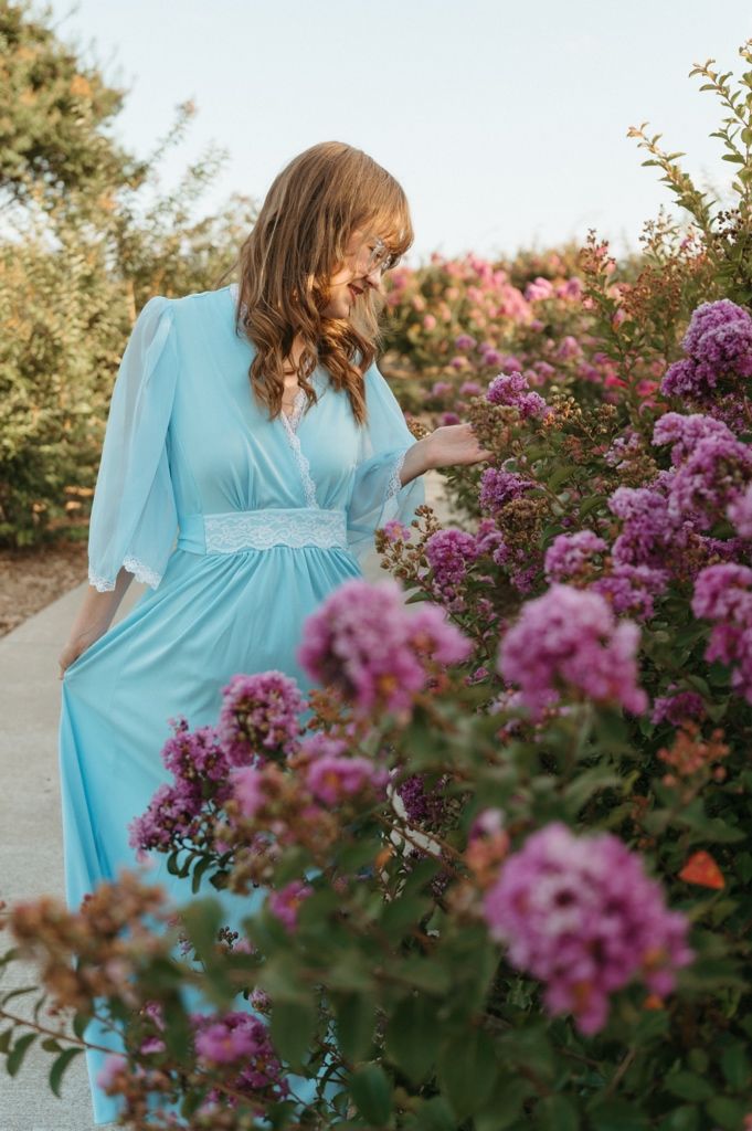 Young woman in light blue chiffon dress gently touches pink flowers on garden bush.