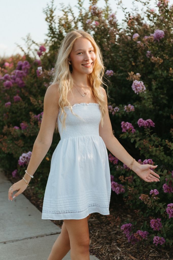 Smiling blonde woman in a white strapless smocked mini dress posing outdoors near blooming pink flower bushes.