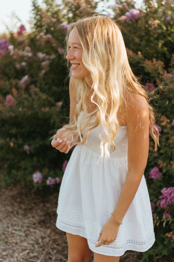 Smiling blonde girl in white sundress twirling among pink flowering bushes in golden hour light.