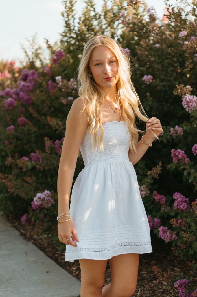 Young woman in white sundress posing before blooming pink flowers in golden hour light.