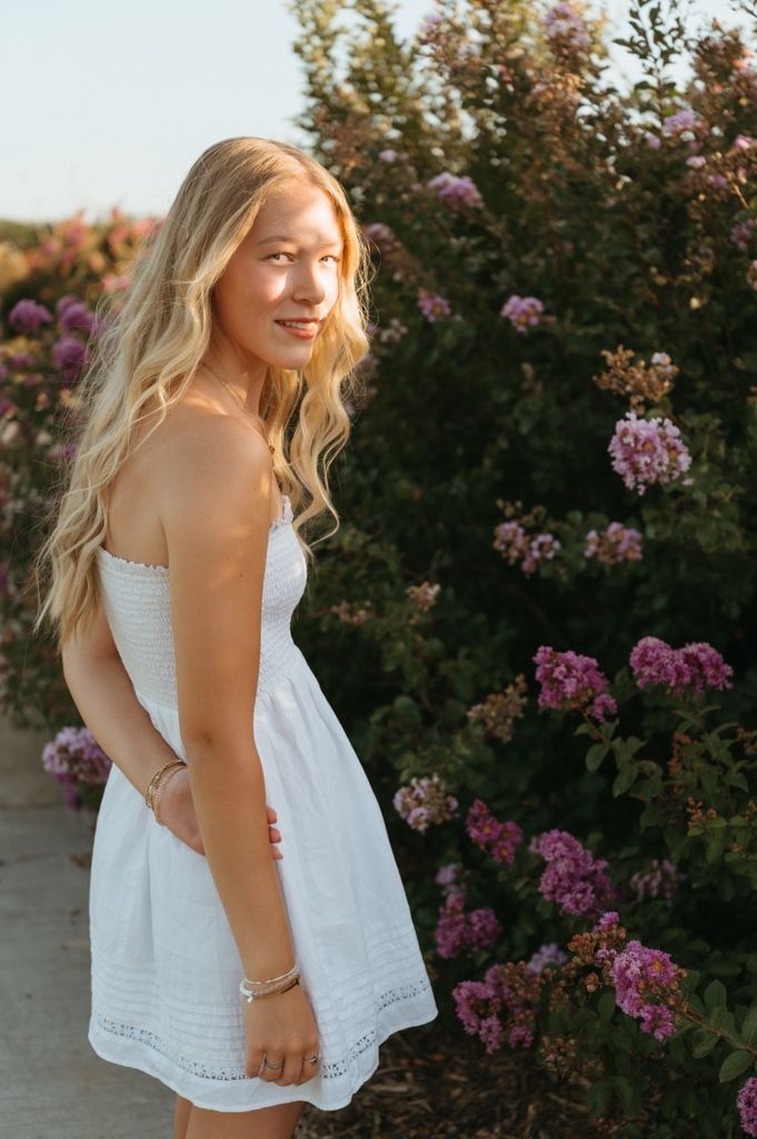 Young blonde woman in white strapless dress posing near blooming pink flowers in golden hour light.