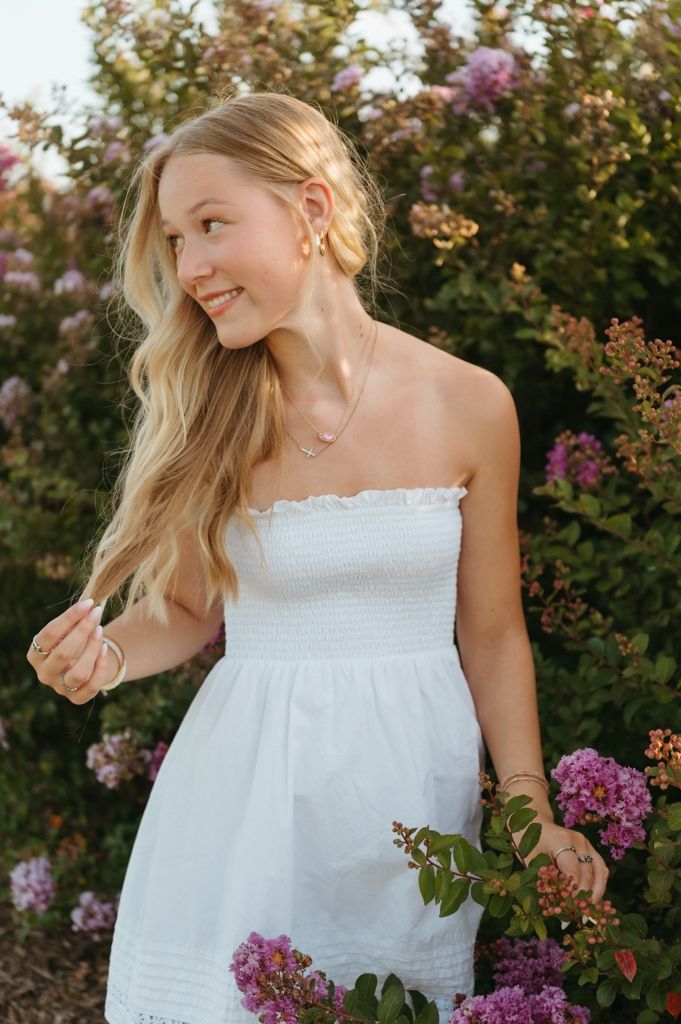 Smiling blonde woman in white strapless smocked dress standing among pink flowering bushes outdoors.