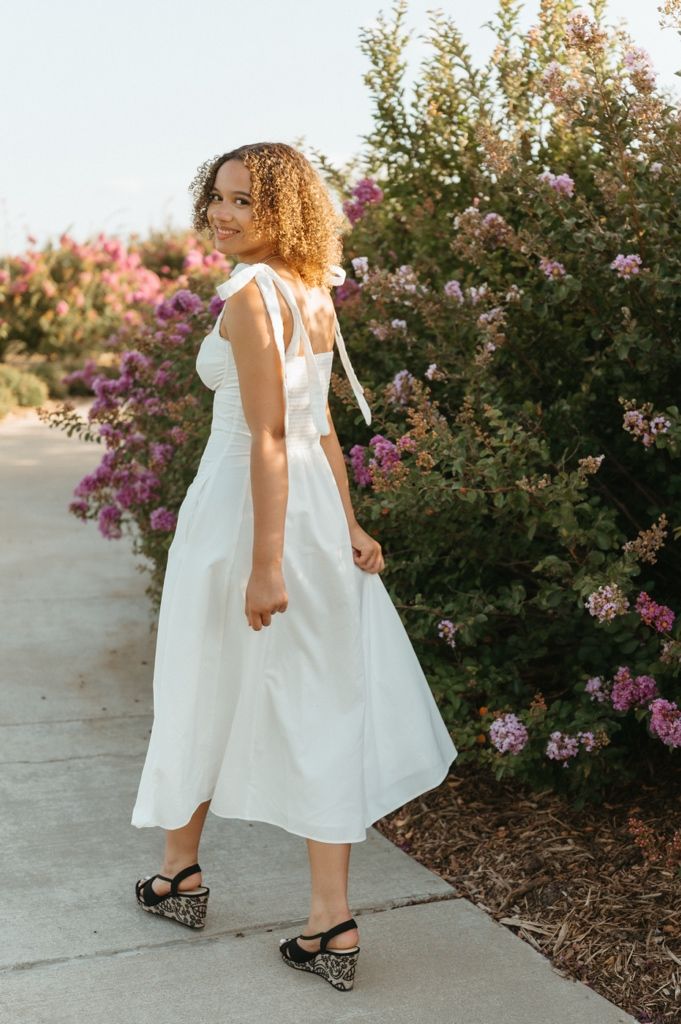 Woman in white midi dress with shoulder ties and black floral flats, posing among blooming pink bushes outdoors.