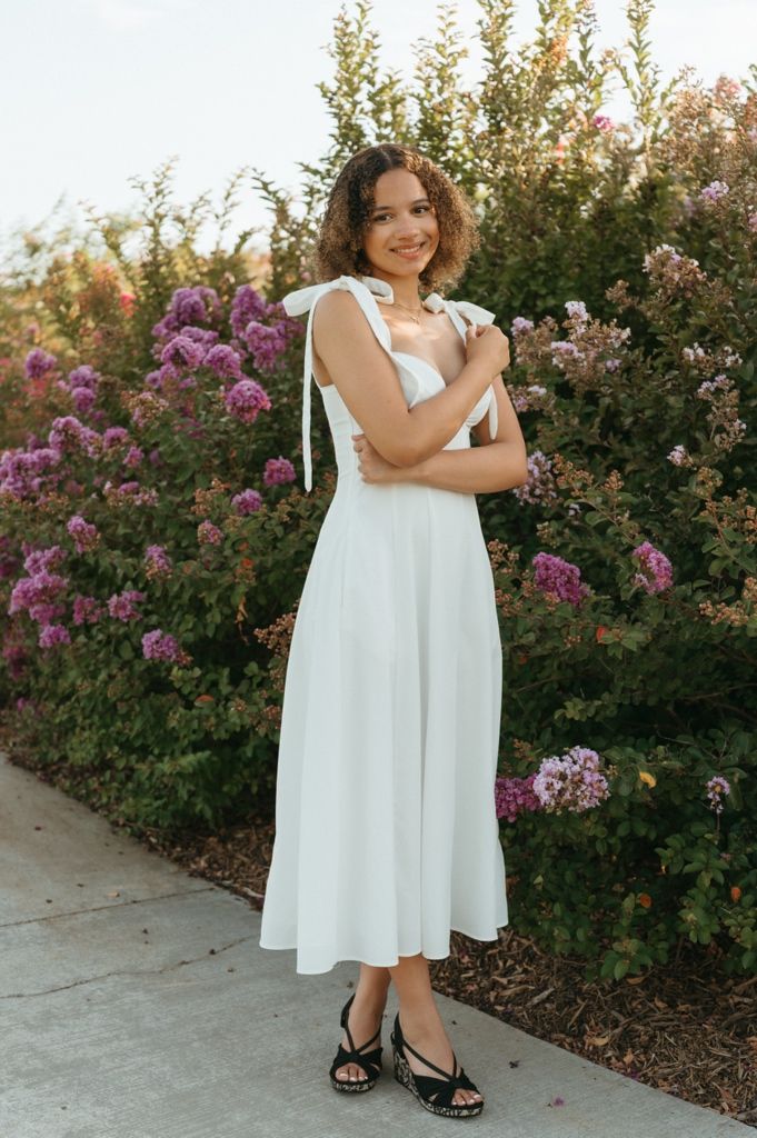 Woman in white midi dress with tie shoulders poses on garden path surrounded by blooming pink flowers.