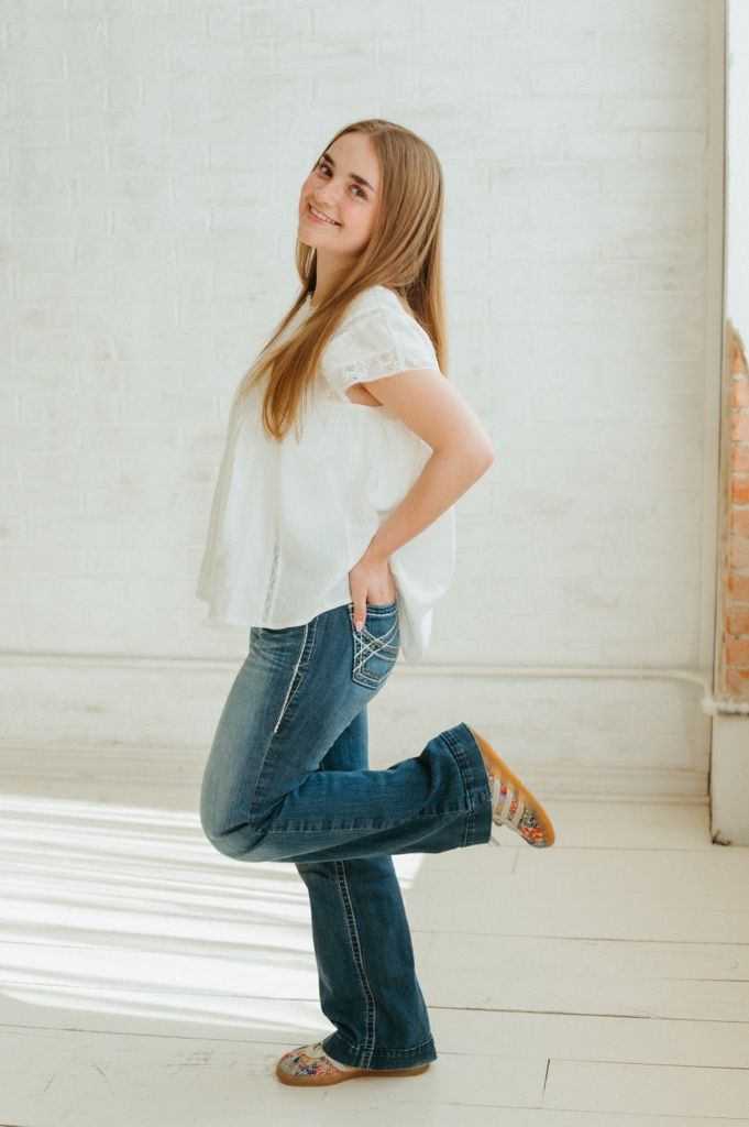 Young woman in white lace top and flare jeans poses playfully in bright white studio.