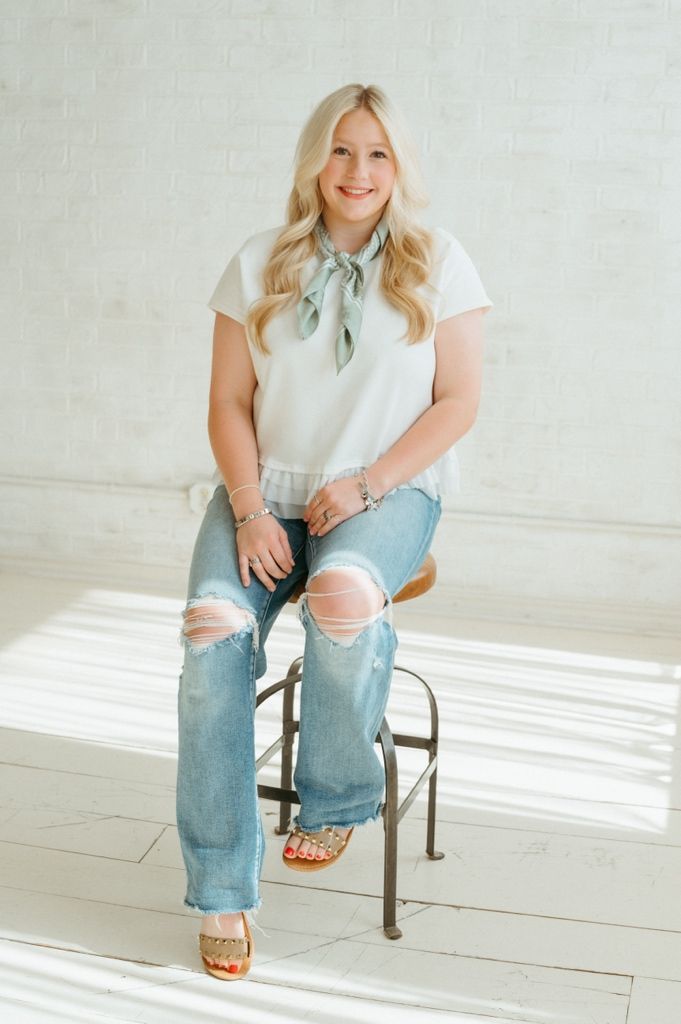 Smiling blonde woman in white top with sage bow and ripped jeans seated on stool by white brick wall.