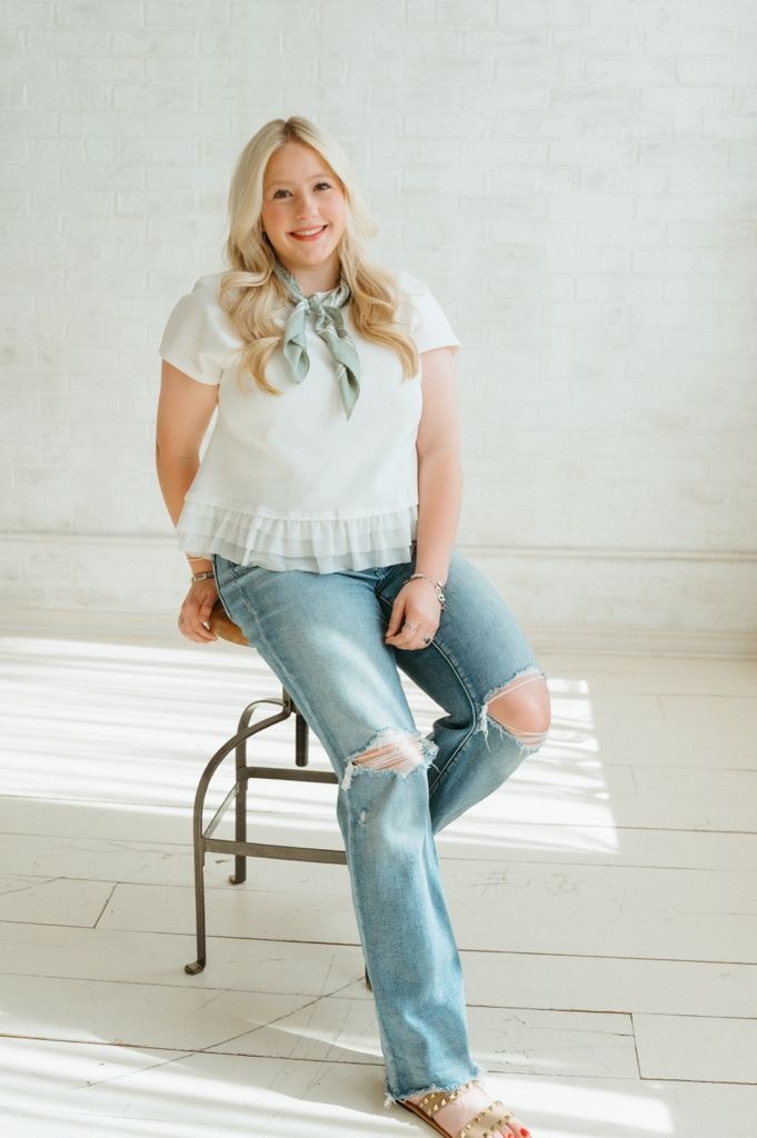Blonde woman in white ruffle top with green bow scarf sits on stool in airy studio.