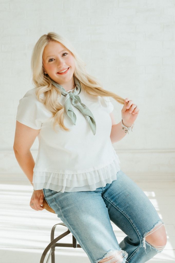 Smiling blonde woman sitting on stool wearing white ruffled top, sage green neck scarf, and distressed jeans.