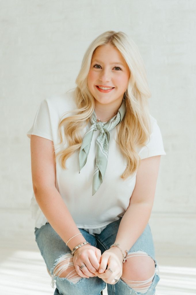 Smiling blonde woman in white top with sage green neck scarf and distressed jeans, sitting casually in bright studio.